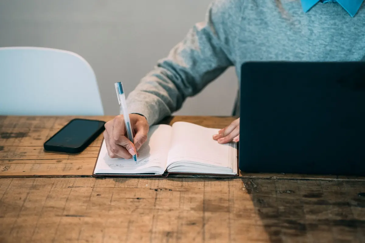 person writing notes in a notebook beside a laptop at a wooden desk