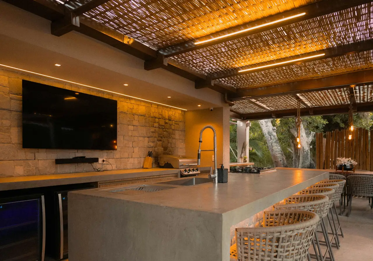 covered outdoor kitchen with a long concrete island, bar stools and a woven shade roof lit warm at dusk